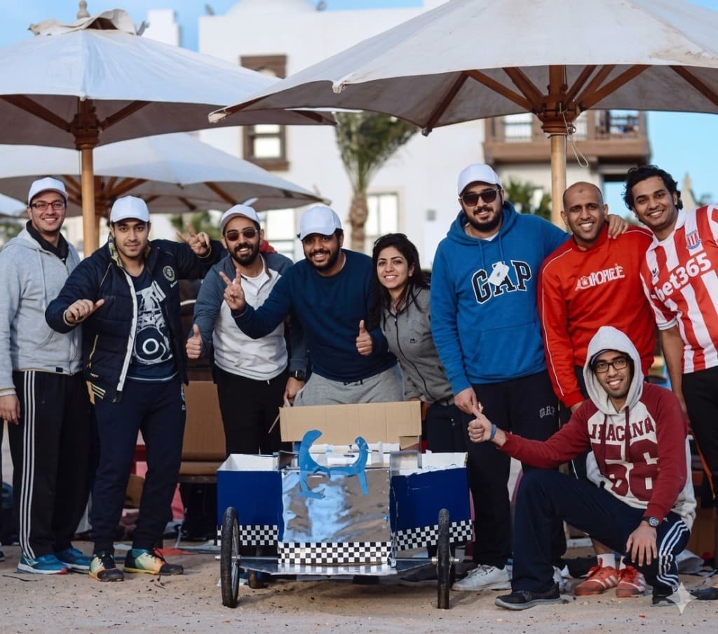 Smiling team members posing with a handmade blue and silver soapbox-style cart on sand under patio umbrellas.