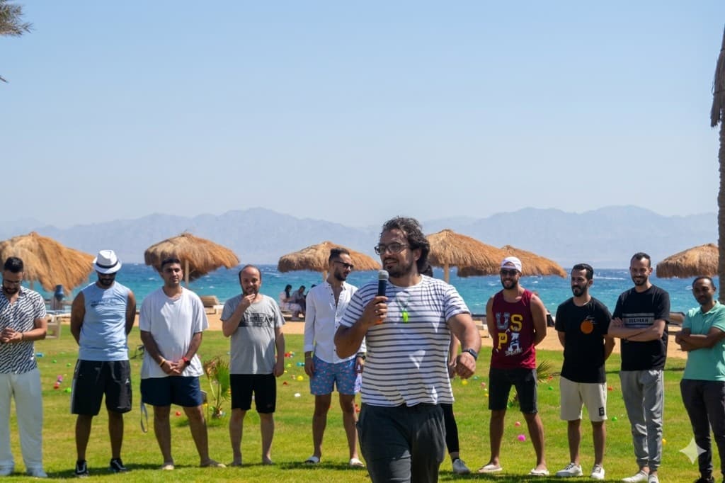Facilitator with a microphone addressing participants on a green lawn by the sea at a sunny beach-side venue.